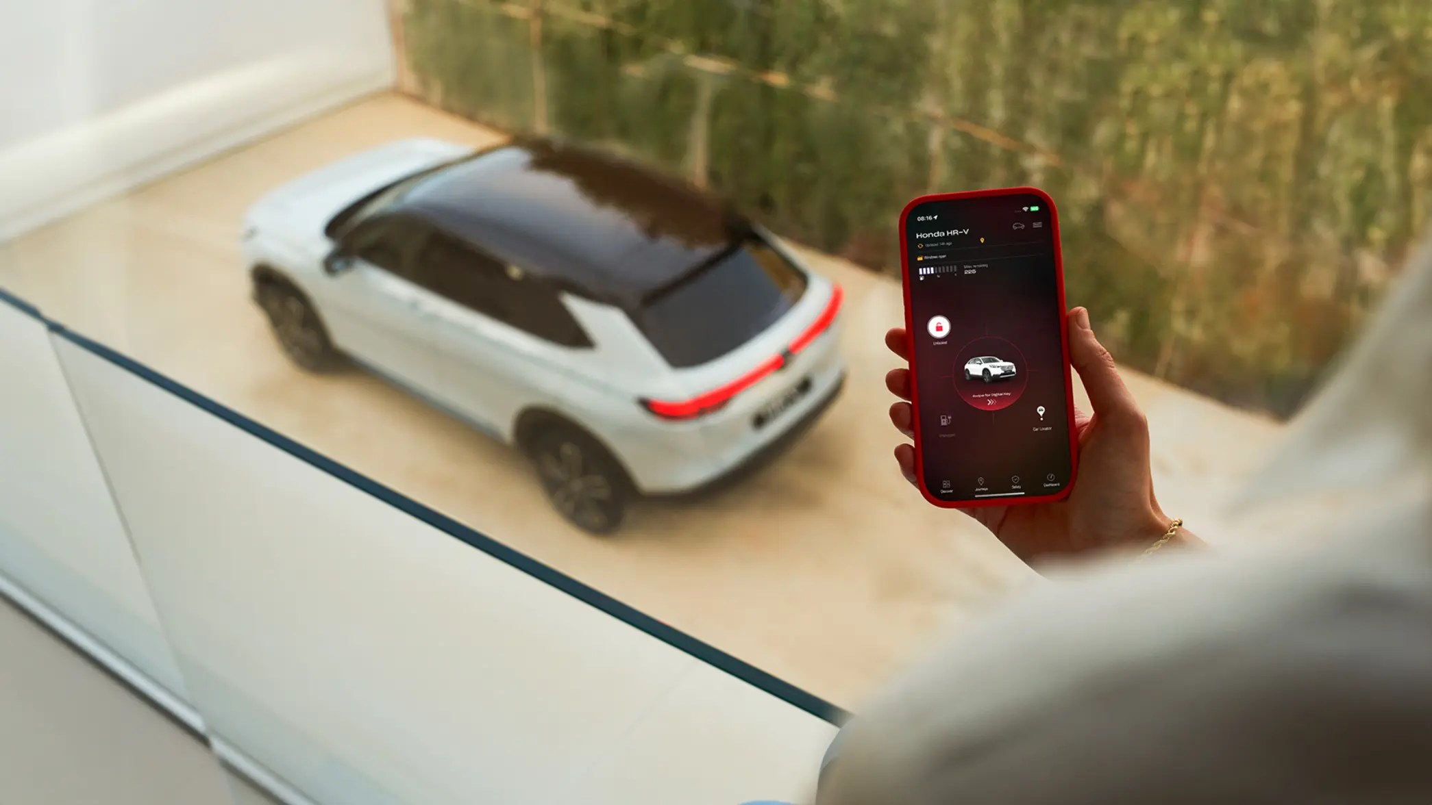 Detailný záber osoby s telefónom, ktorá používa aplikáciu My Honda +, pričom vozidlo Honda HR-V vidieť za oknom